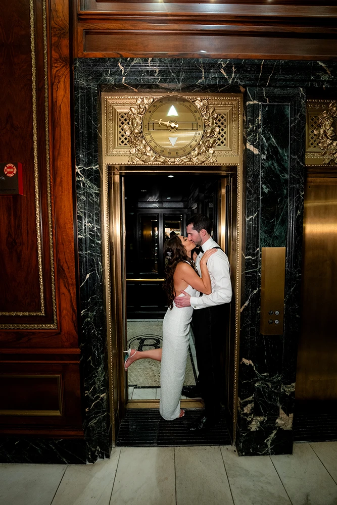 couple kissing and blocking the door of the elevator surrounded by gold elements and lots of marble details